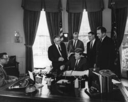 President John F. Kennedy (seated at desk) signs an amendment to the Soil Conservation and Domestic Allotment Act to limit the drainage of wetlands. Standing behind President Kennedy (L-R): Representative Lester R. Johnson (Wisconsin); Assistant Secretary of Agriculture, John P. Duncan, Jr.; Representative Henry S. Reuss (Wisconsin); Senator Eugene J. McCarthy (Minnesota). An unidentified stenographer sits at far left. Oval Office, White House, Washington, D.C. 1962. Public Domain.